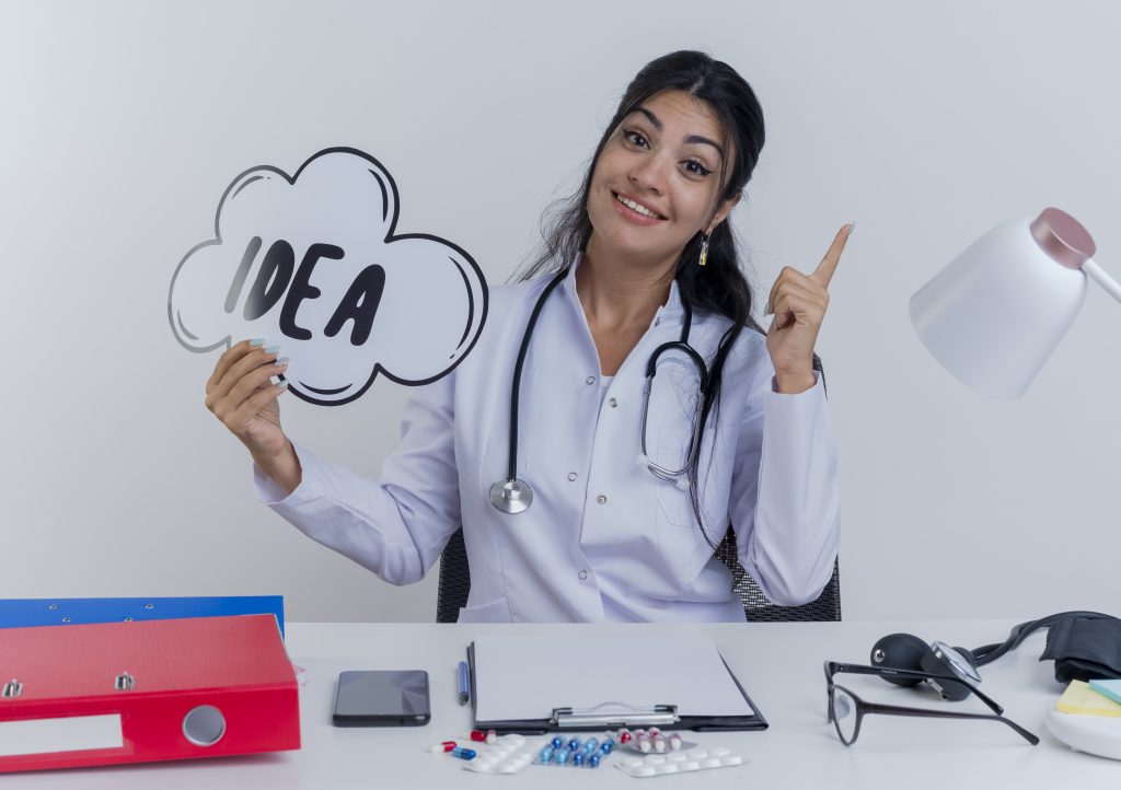 impressed young female doctor wearing medical robe and stethoscope sitting at desk with medical tools holding idea bubble looking at camera smiling and raising finger isolated on white background